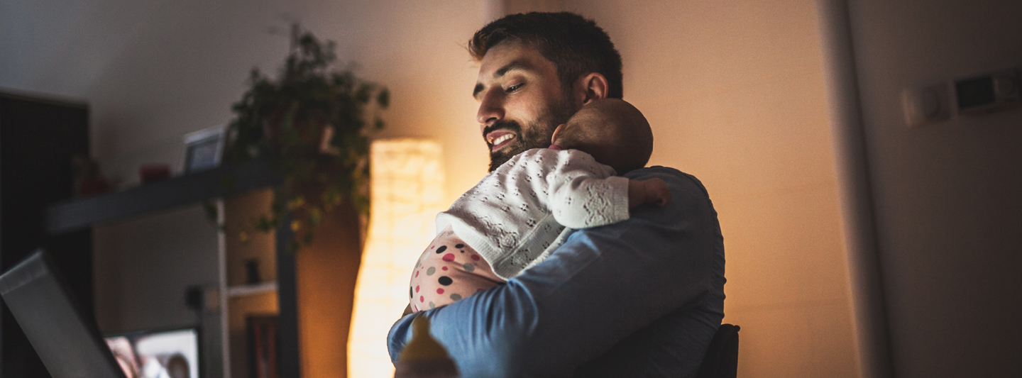 Young father holding sleeping baby while on laptop